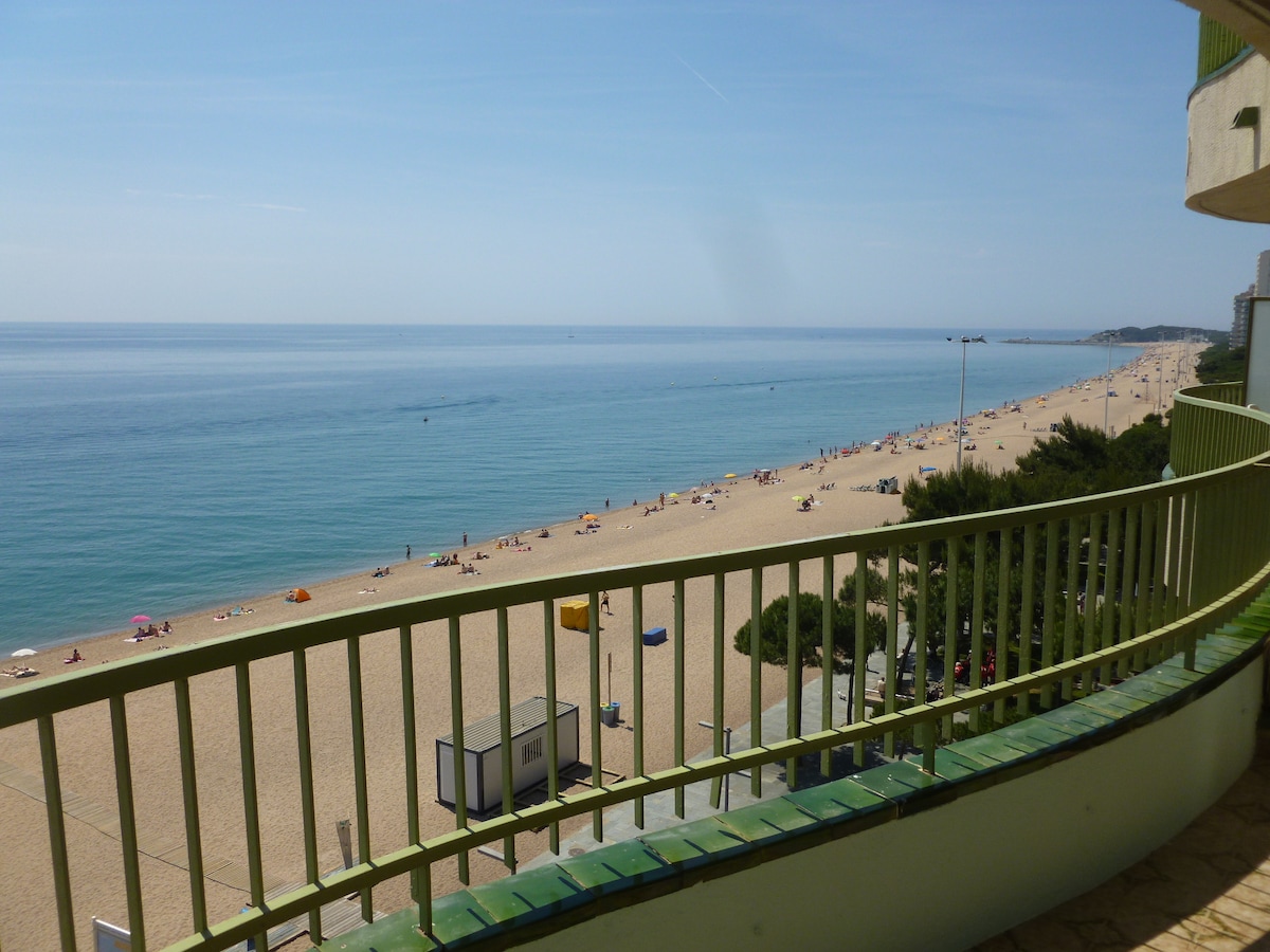 A spacious terrace offers unobstructed views of the sandy beach and calm blue sea. Sunbathers can be seen along the shoreline, with a few parasols scattered on the sand. The inviting horizon blends into the clear sky, creating a serene atmosphere.