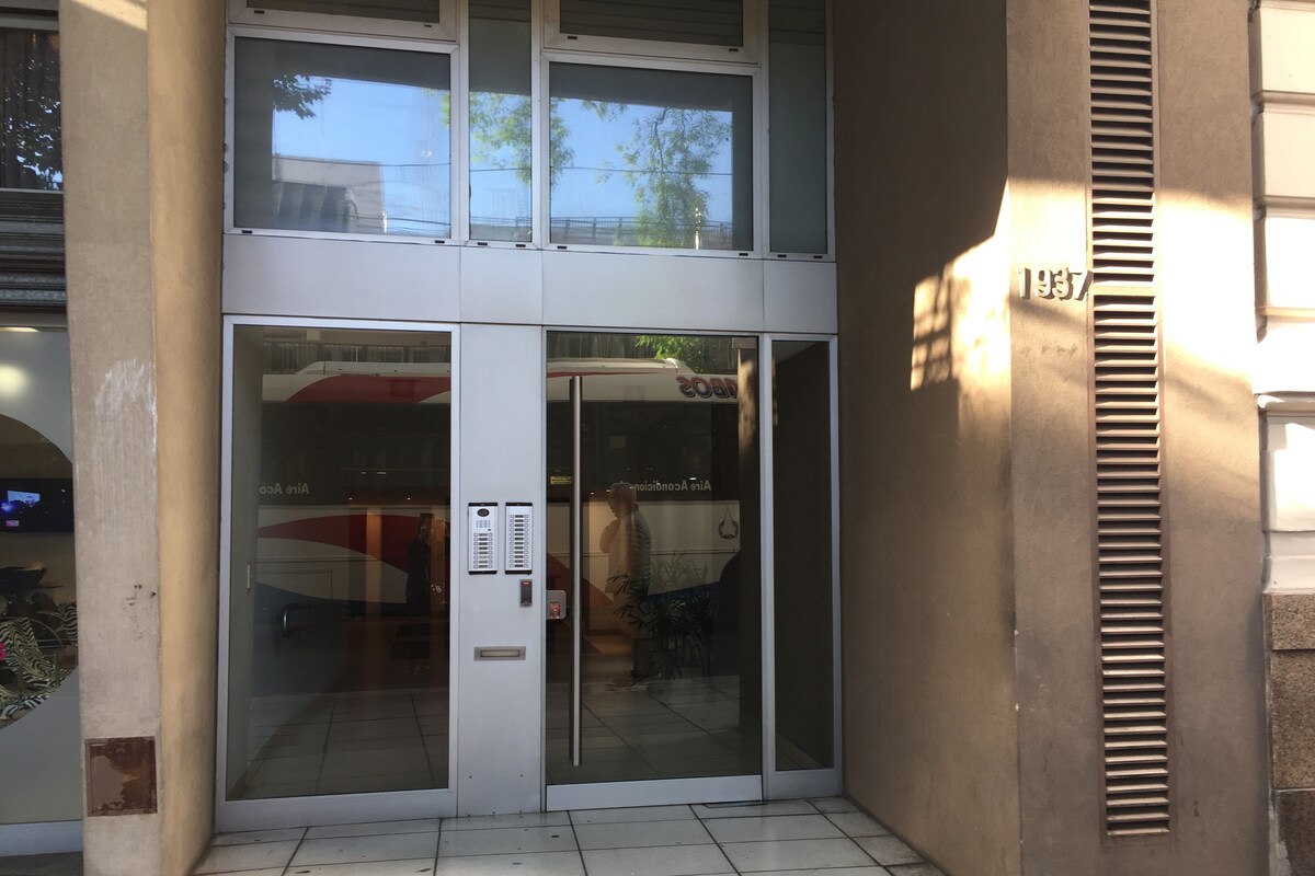 The entrance of the building is presented with large glass doors, allowing natural light to illuminate the lobby area. A minimalist design is featured, with a beige exterior and vertical slats beside the entrance. A small intercom panel is situated adjacent to the doors.