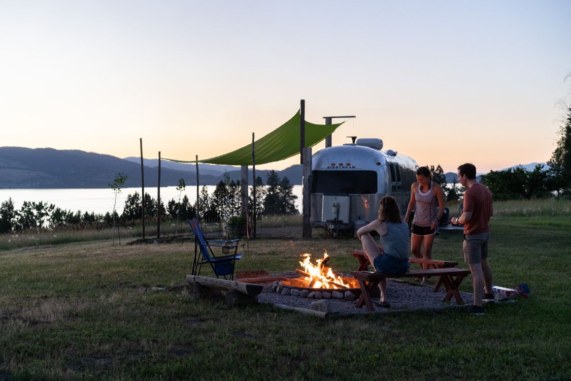 A cozy fire pit arrangement is shown, surrounded by seating and green shade cloth. In the background, a vintage airstream reflects the colors of the sunset overlooking Flathead Lake. Trees and mountains provide a serene backdrop to the scene.