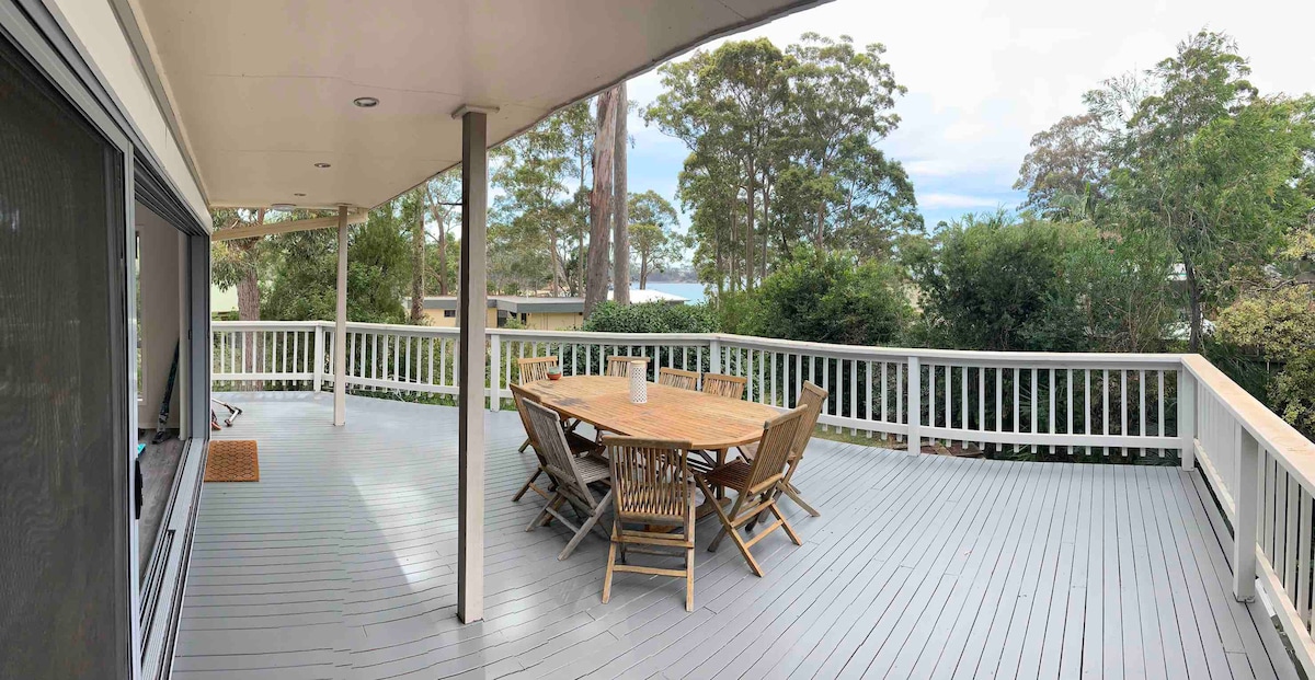 A spacious outdoor deck features a large wooden dining table surrounded by six chairs. The area is shaded by an overhang and offers views of lush greenery. Sunlight filters through the trees, creating a serene atmosphere ideal for gatherings.