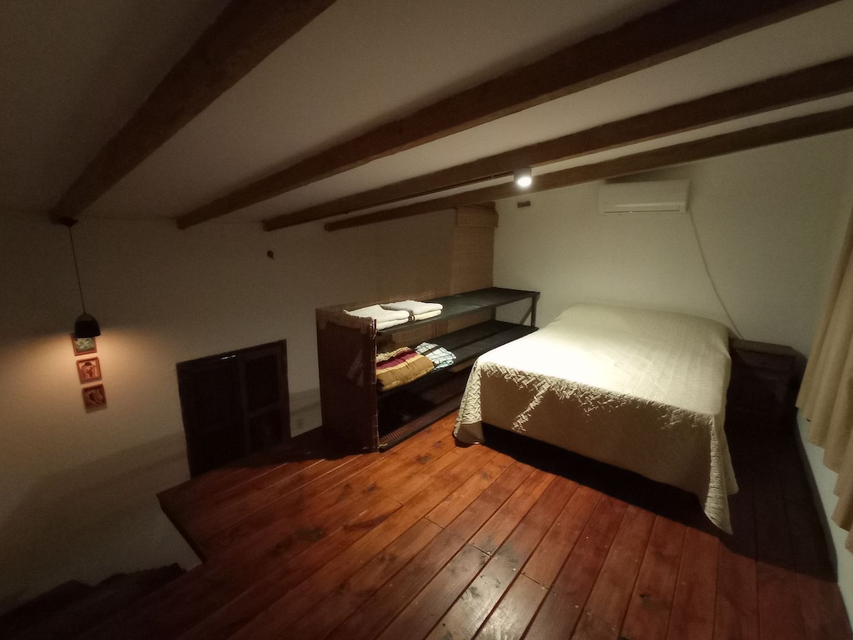 An inviting sleeping area features a spacious bed dressed in a light-colored cover. A wooden shelf displays neatly folded linens, while natural wood beams add warmth to the ceiling. A window is fitted with light-filtering curtains, complementing the cozy ambiance.