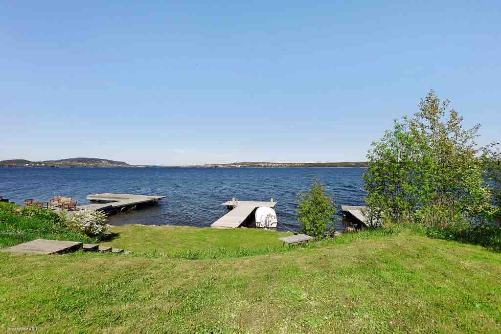 A serene lakeside scene features two wooden docks extending into the calm water. Lush green grass covers the foreground, interspersed with shrubs and a young tree, while clear blue skies reflect above the tranquil lake.