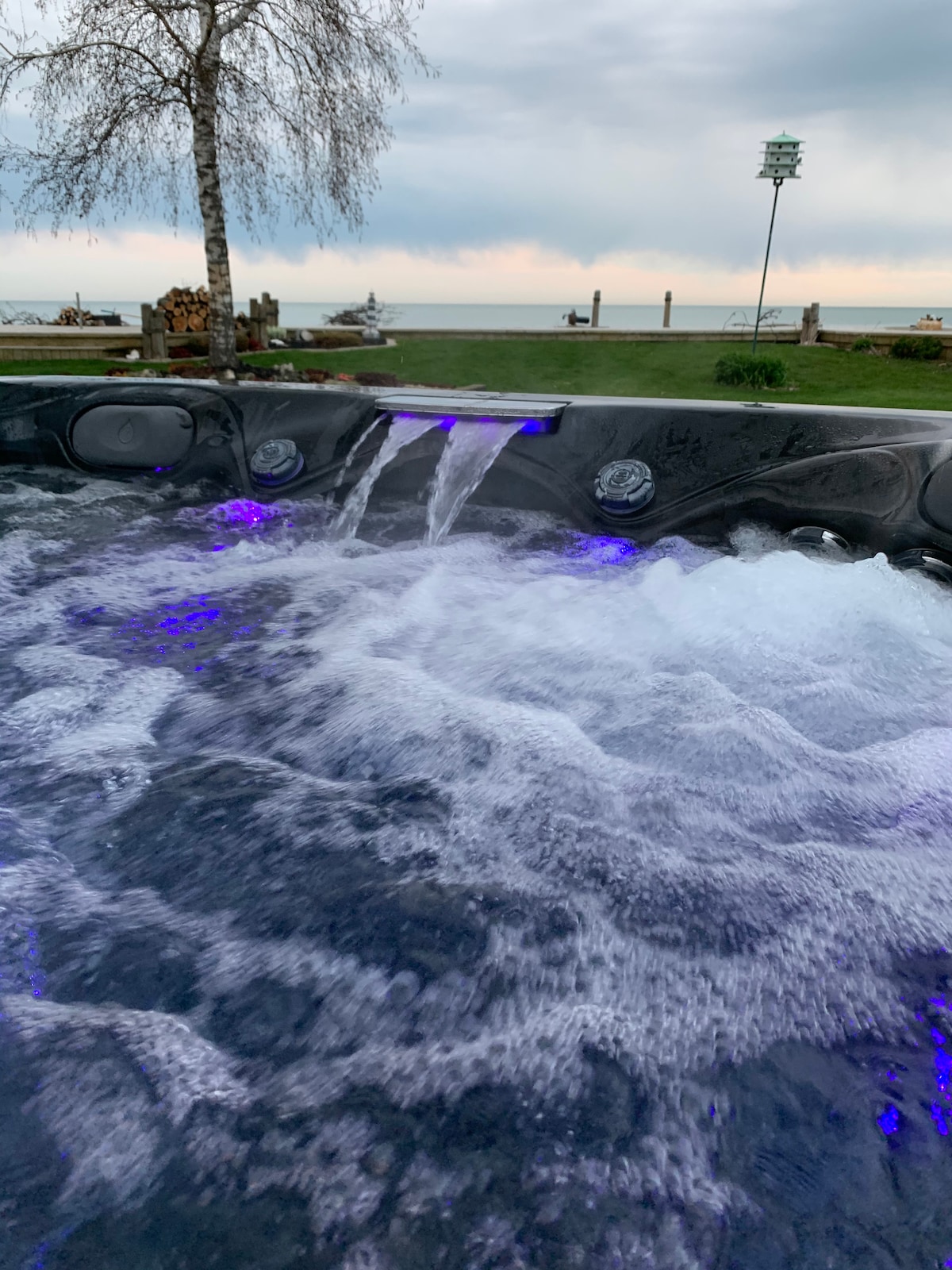 A hot tub with bubbling water is shown, featuring illuminated jets in soft purple hues. The backdrop includes a scenic view of the lake and grassy shoreline, with a glimpse of distant boats. Natural elements are present, such as a nearby tree and stacked firewood.