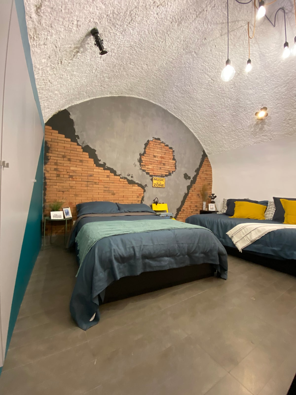 A spacious bedroom featuring a barrel-vaulted ceiling and a striking accent wall of exposed brick. The room includes a large bed with navy bedding and decorative yellow and grey cushions. Warm lighting is provided by pendant lights hanging from the ceiling.