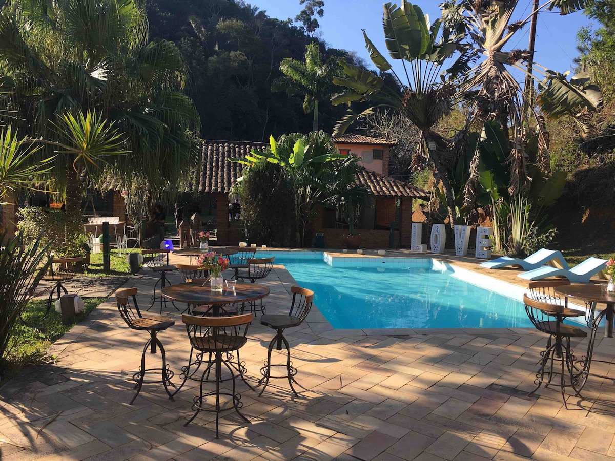 A clear blue pool is surrounded by a paved patio with metal tables and chairs. Lush tropical plants provide a serene backdrop, with a charming building visible in the distance. A large decorative 'LOVE' sign is placed near the pool, adding a welcoming touch.