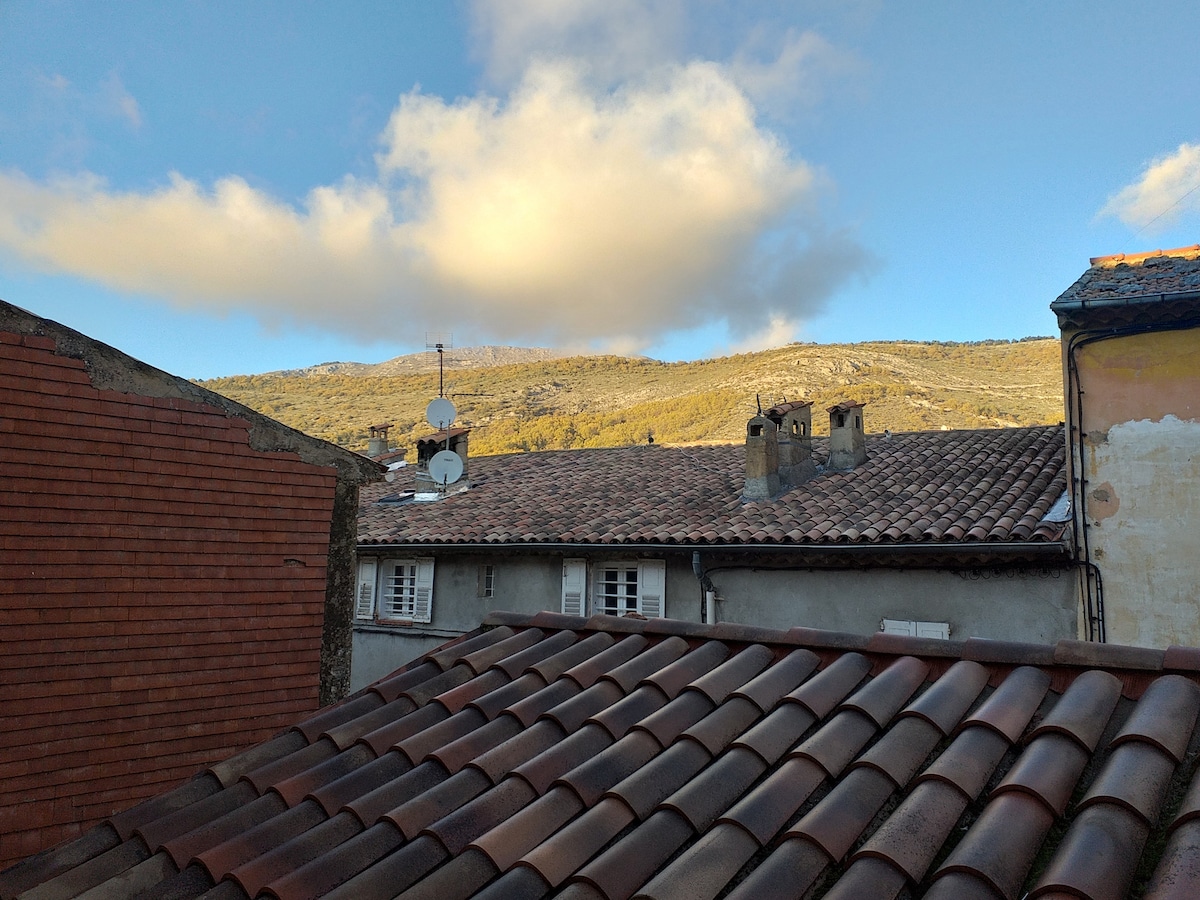 The image captures rooftops of the village, featuring terracotta tiles and stone buildings. A mountain range is visible in the background, under a partly cloudy sky. The scene offers a serene view of the surroundings, indicative of the area's natural landscape.