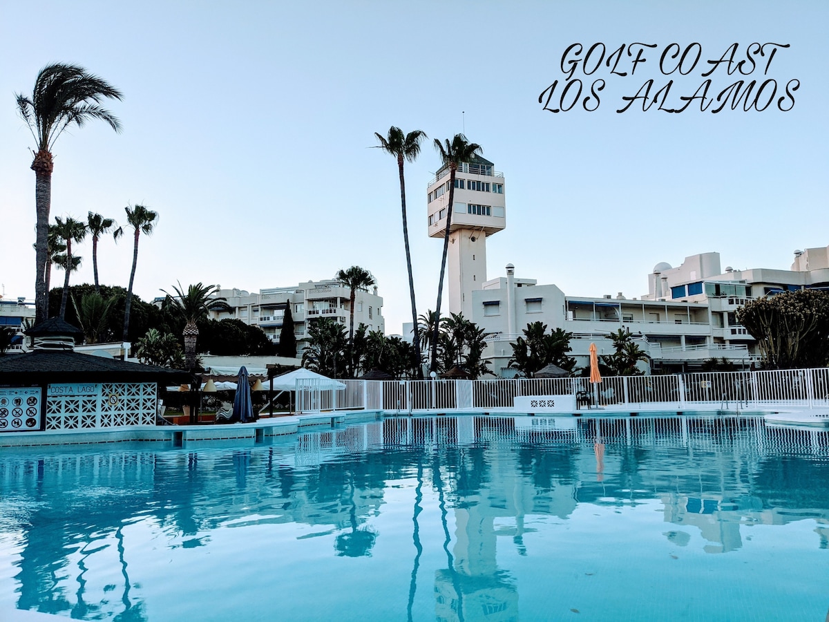 A serene swimming pool reflects the clear sky, surrounded by palm trees and well-maintained gardens. The buildings in the background house various amenities and accommodations. A tower stands prominently, adding architectural interest to the scene.