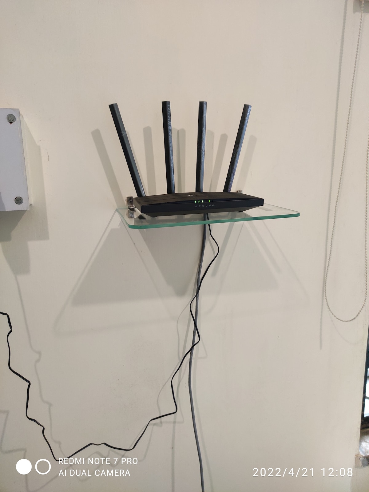 A wireless router is mounted on a transparent glass shelf against a light-colored wall. Five antennas extend upwards from the device, and a cable is connected to it, trailing downwards. The setup provides connectivity within the apartment.