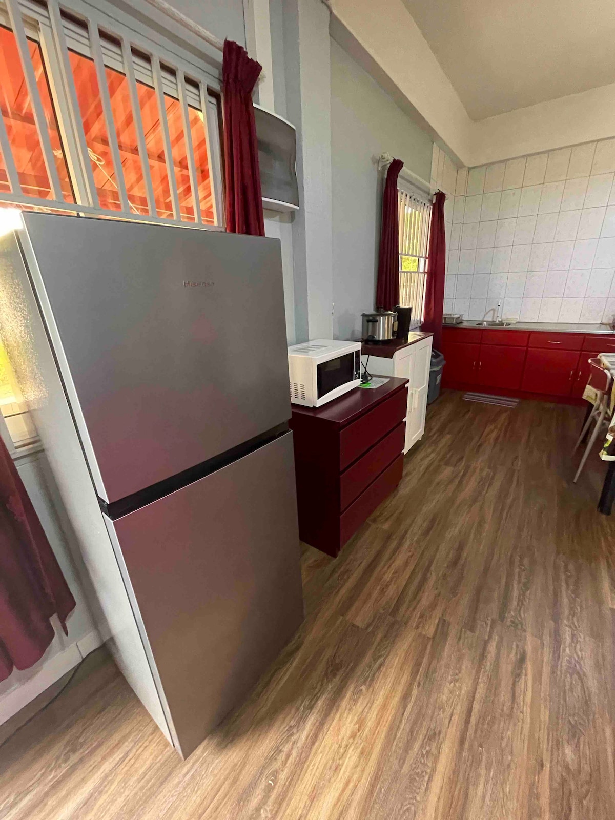 The kitchen area features a silver refrigerator alongside a microwave and a small red cabinet. Light wooden flooring is present, complemented by red cabinetry in the rear. Natural light filters through the window, partially covered by burgundy curtains.