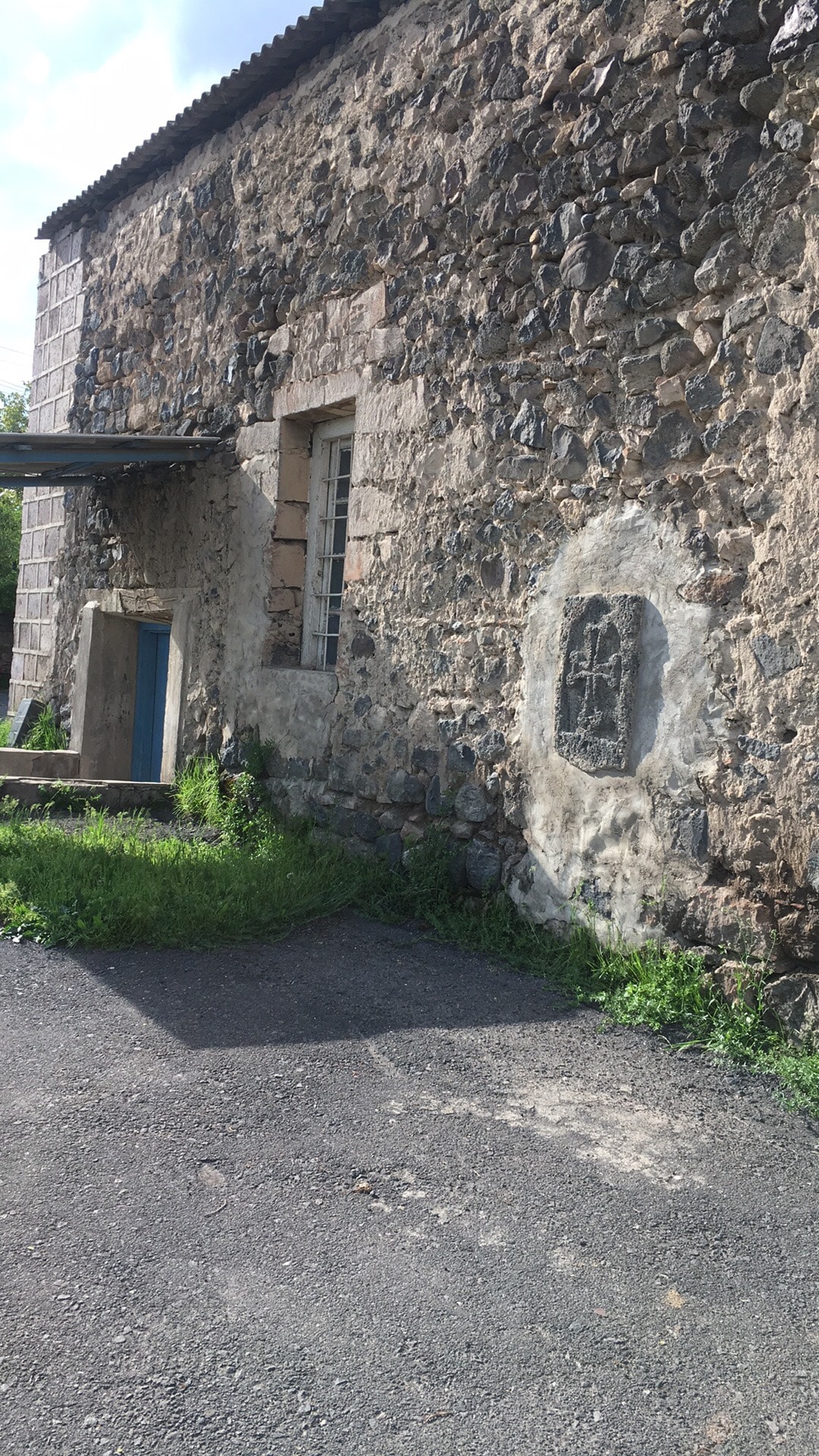 The exterior of a rustic stone building is visible, showcasing a textured stone facade with patches of greenery at its base. The entrance features a wooden door, accompanied by a window partially obscured by the surrounding wall. Sunlight casts shadows, highlighting the structure's age and charm.