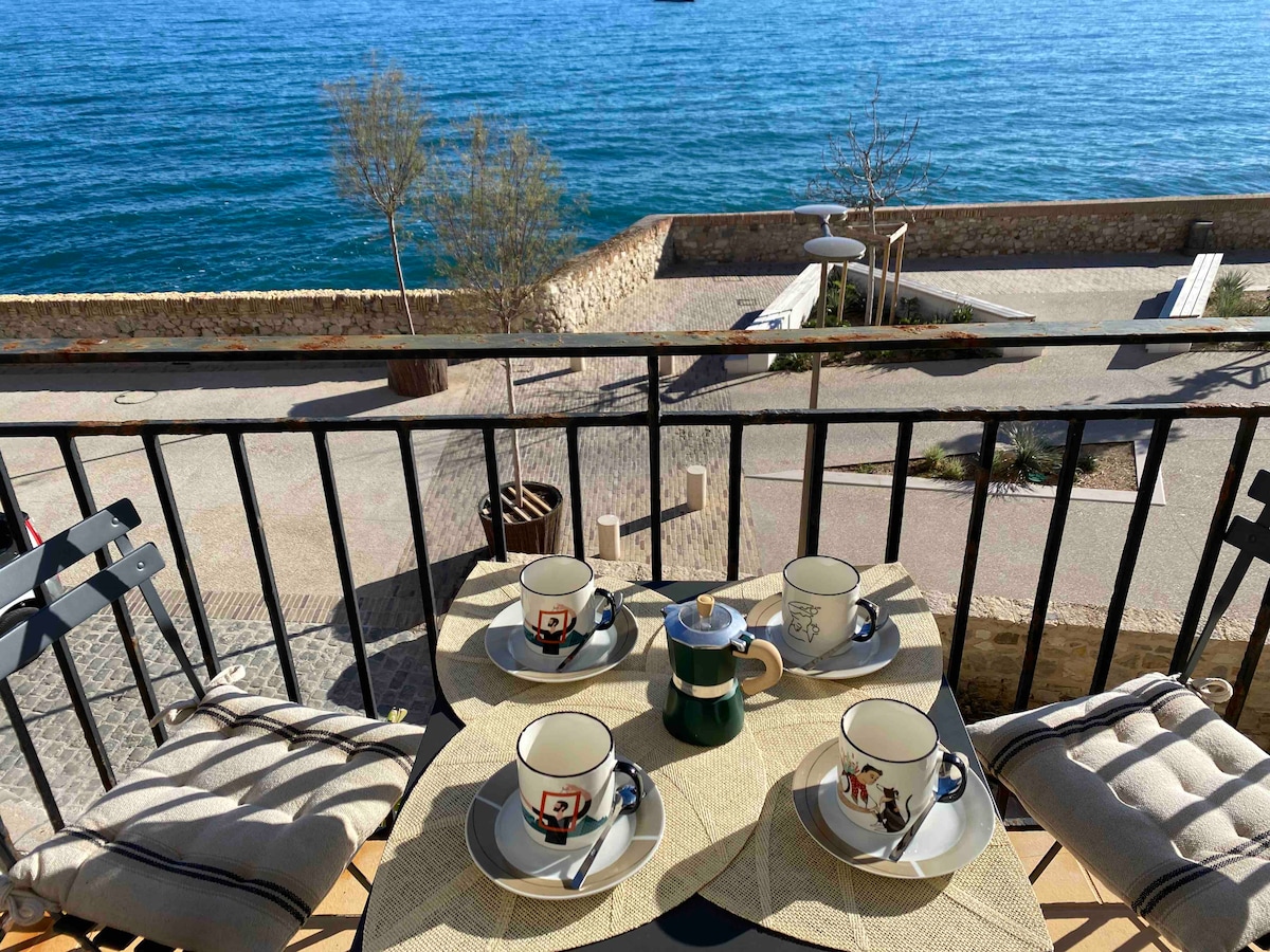 A cozy balcony view features a small round table set for four, with cups and a coffee pot arranged on a beige placemat. The backdrop showcases a serene sea view, with calm waters and clear skies. Nearby, a tree and a stone wall are visible.