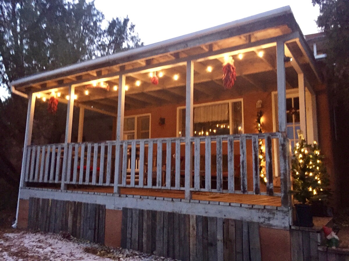 A charming private porch is showcased, illuminated by string lights and adorned with decorative elements. The wooden railing adds warmth, while the surrounding greenery complements the cozy ambiance. A small tree is featured, enhancing the inviting atmosphere for outdoor relaxation.