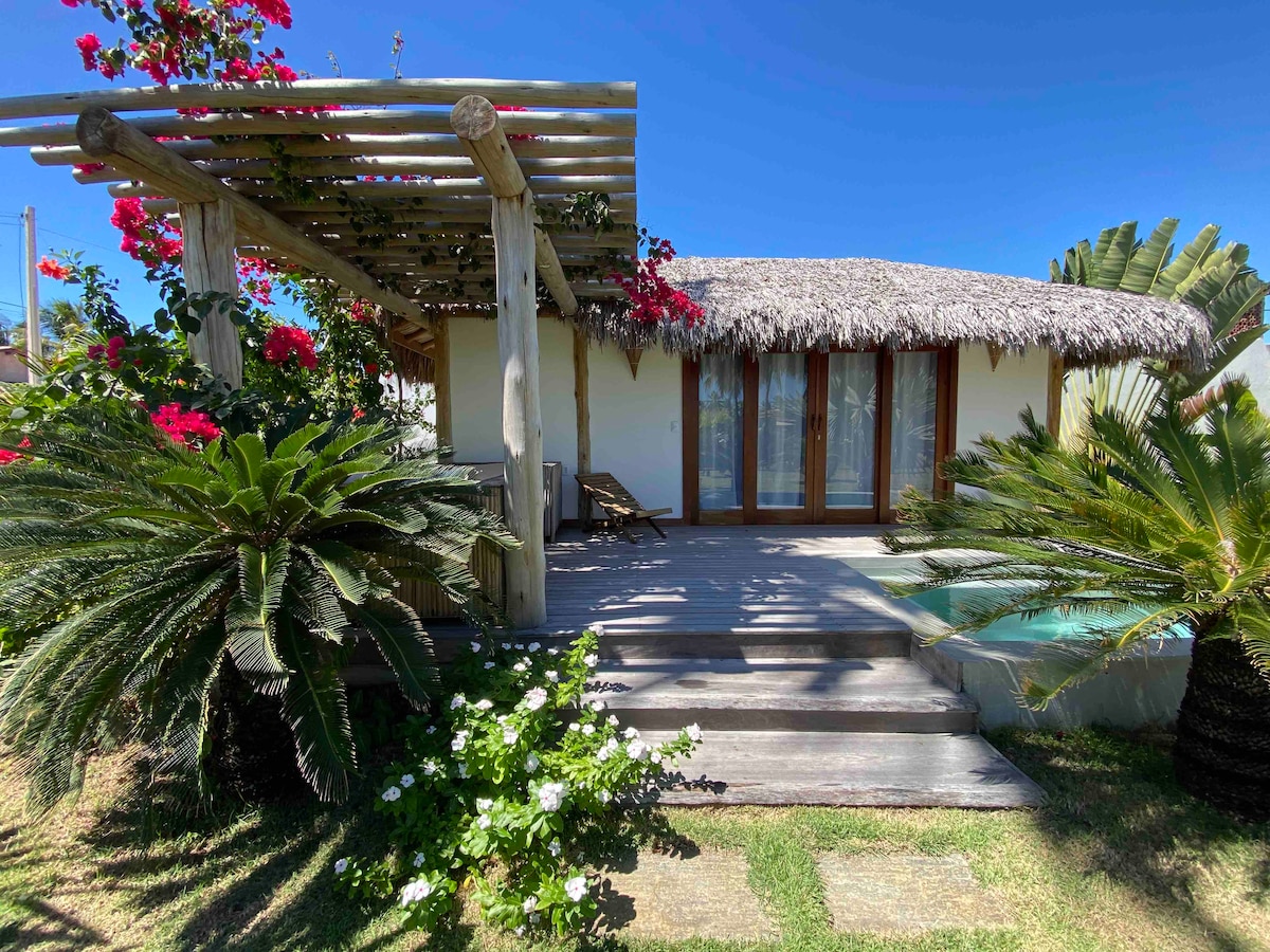 A bungalow with a thatched roof is surrounded by lush greenery and colorful flowers. A wooden deck leads to large glass doors. The serene atmosphere is enhanced by a private pool visible to the side, framed by tropical plants.