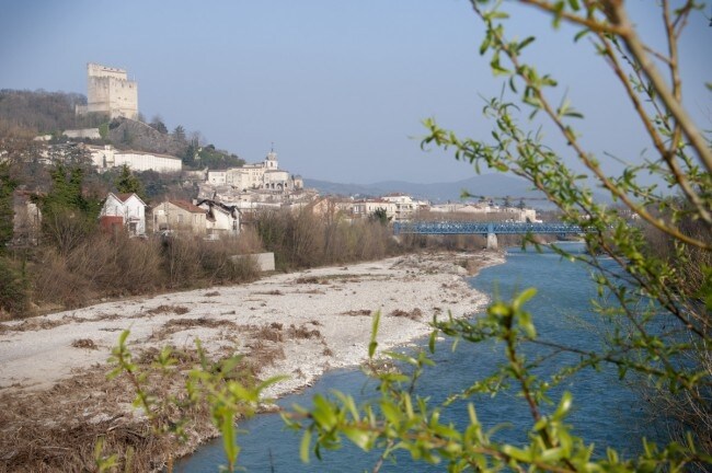 A scenic view of Crest features a historic tower rising on a hilltop, surrounded by a mix of greenery and the flowing Drôme river. The riverside landscape presents pebbles and a clear sky, while the town is visible in the background, highlighting its charming architecture.