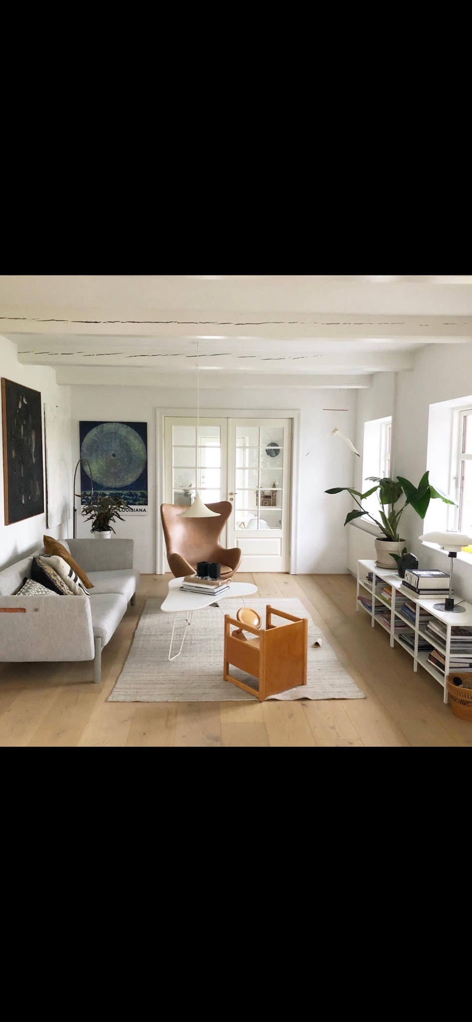 A bright living area features a light gray sofa complemented by decorative pillows. A large wall artwork is present, and a modern chair adds a stylish touch. Natural light filters through multiple windows, illuminating a neutral rug placed beneath a small coffee table.