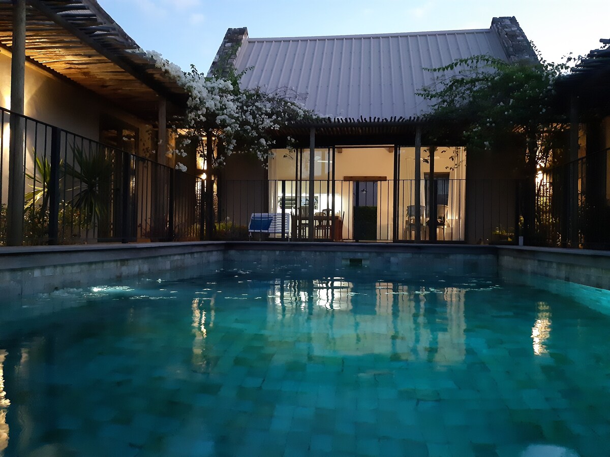 A tranquil magnesium private pool reflects the surrounding architecture, bordered by a secure railing. A spacious patio area is visible through the glass doors, showcasing an outdoor dining setup under a structure accented with greenery and natural materials.
