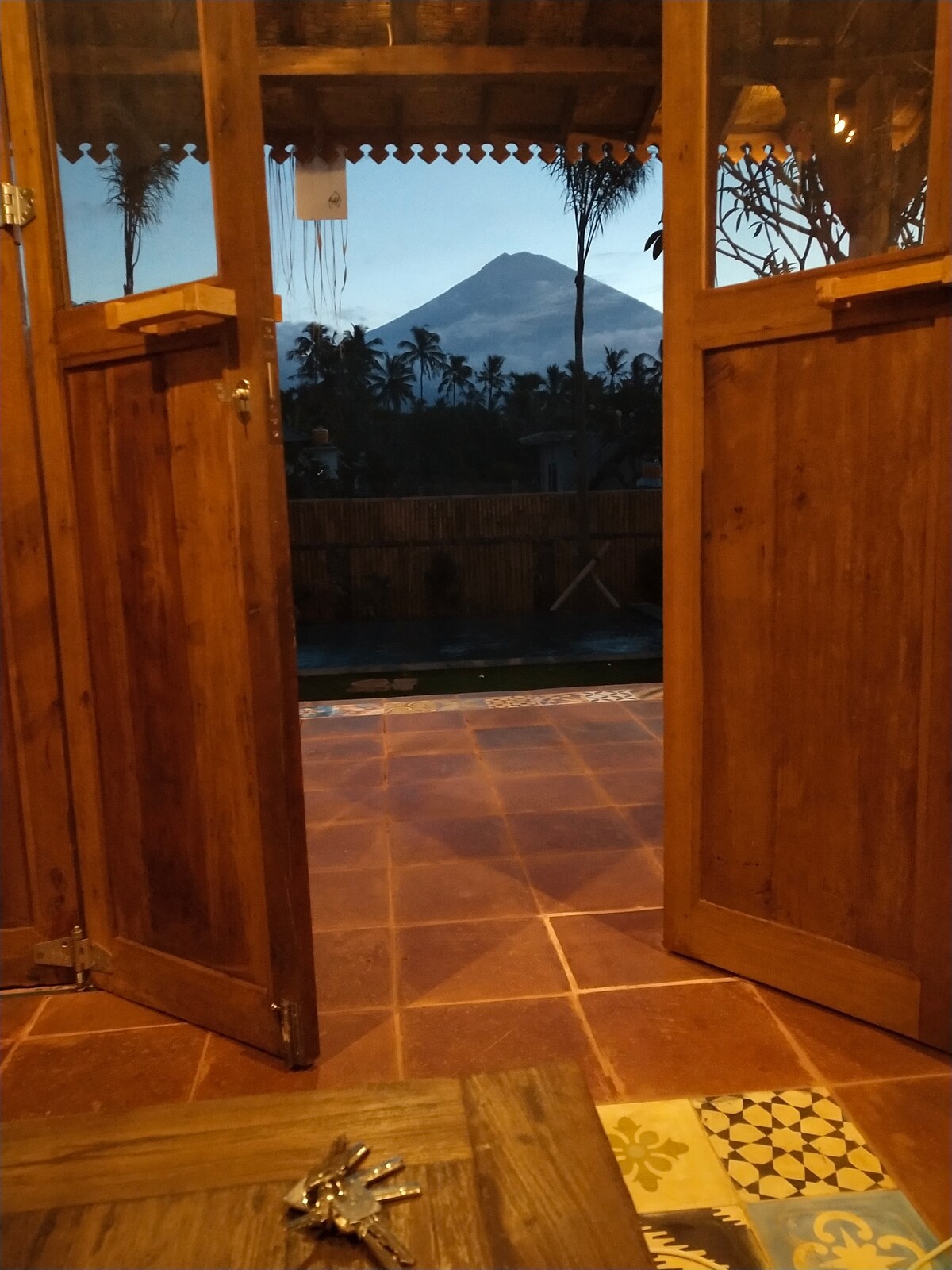 Wooden doors open to reveal an outdoor view with lush palm trees and a mountain in the distance. The floor features colorful tiles, and a serene evening light adds warmth to the scene.