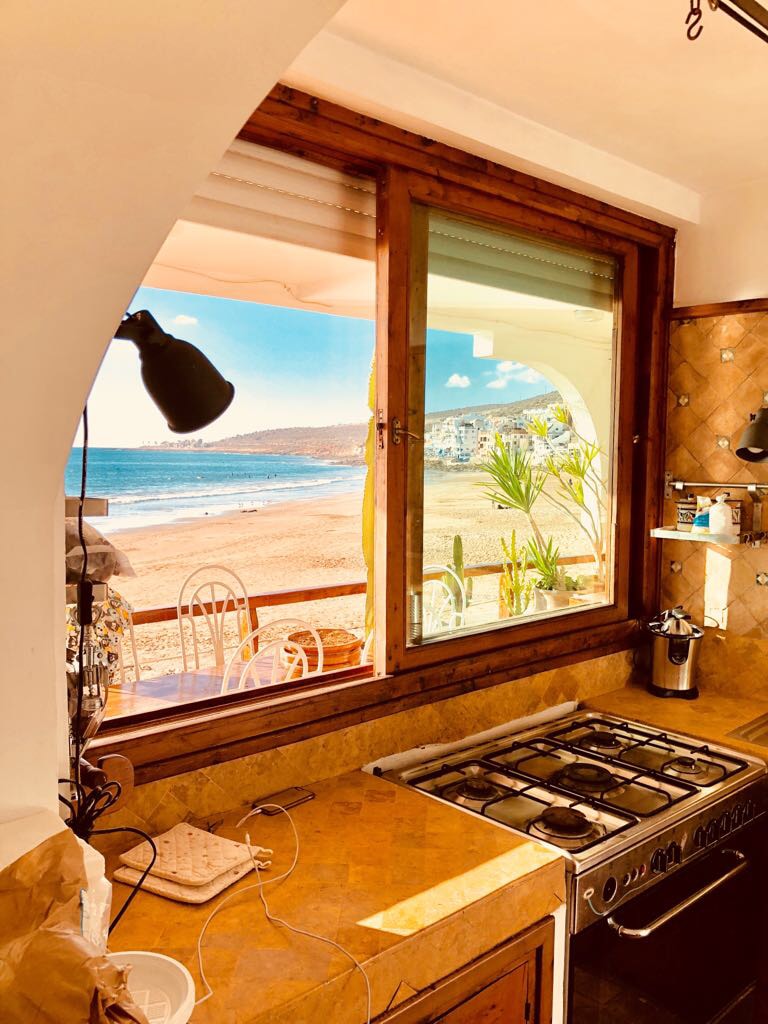 A kitchen area is visible, featuring a gas stove and a large window that frames a view of the sandy beach and ocean. Light from the window illuminates the space, while plants outside add a touch of greenery.