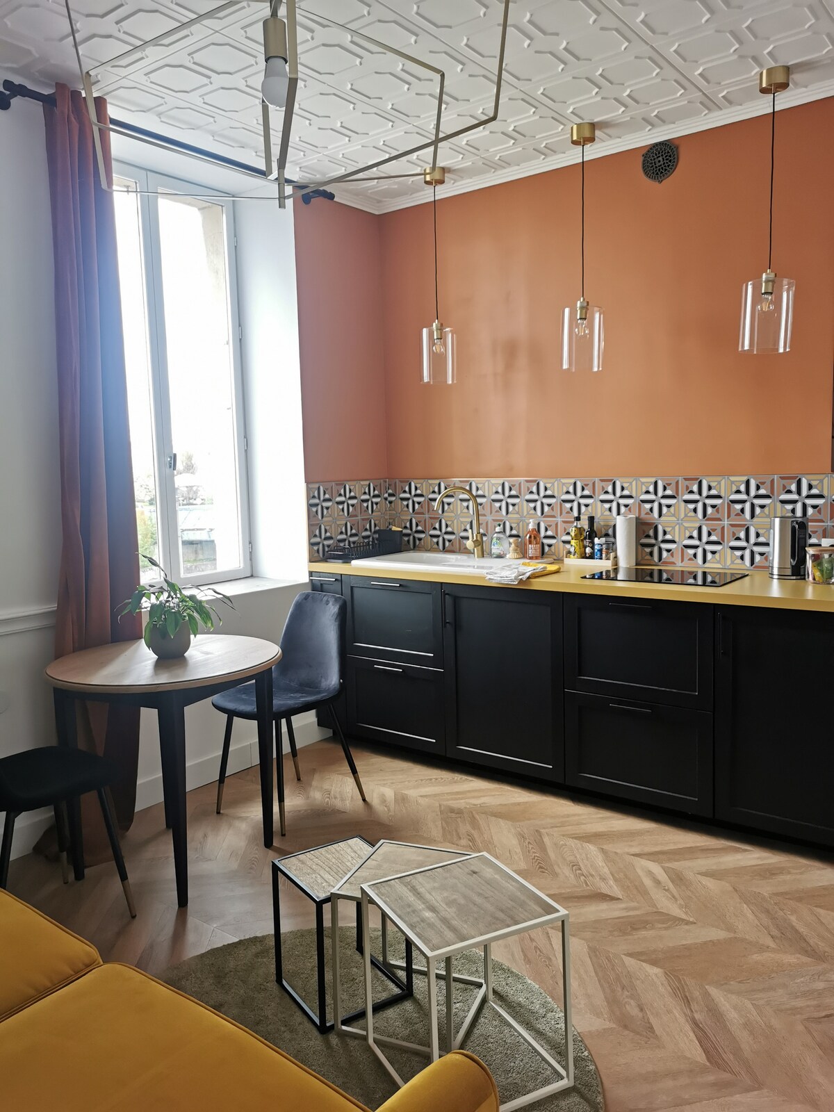 A cozy corner of the apartment features a modern kitchen with dark cabinets and patterned backsplash. A round table with two chairs is positioned beside a large window, allowing natural light to fill the space. Geometric side tables are placed nearby, adding a touch of style.
