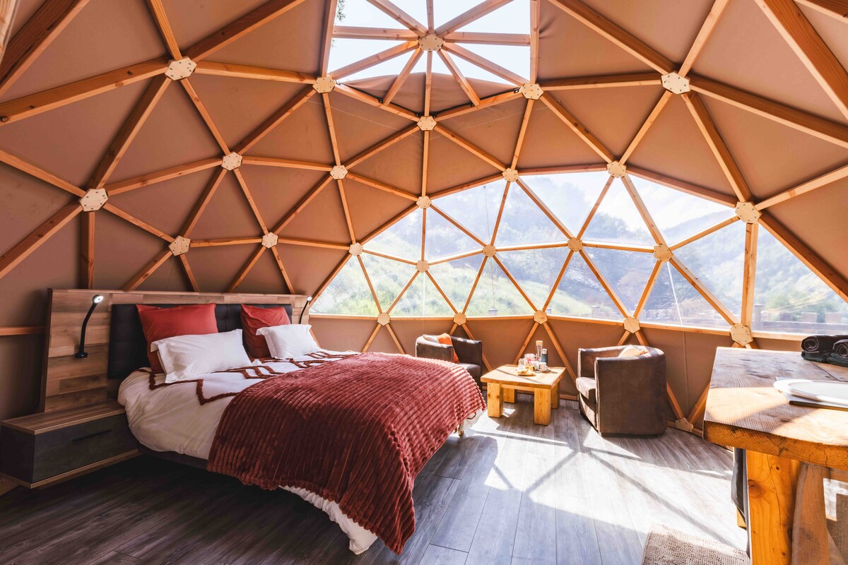 A geodesic dome interior showcases large windows providing panoramic views of the surrounding landscape. A spacious bed is centered, draped with soft bedding and a textured throw. Seating areas with armchairs and a small table create a cozy atmosphere, complemented by natural wood accents throughout.