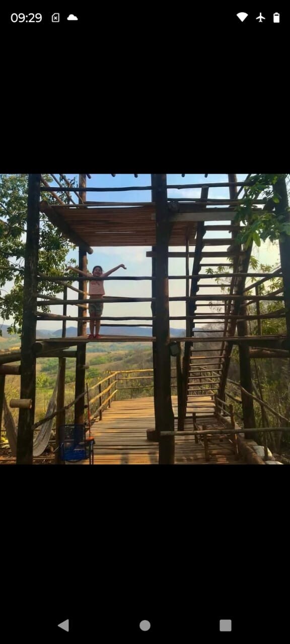 A wooden structure featuring a multi-level design showcases open spaces supported by sturdy posts and railings. Green trees frame the scene, and a person stands on the upper level, extending an arm towards the expansive view of the valley and surrounding nature.