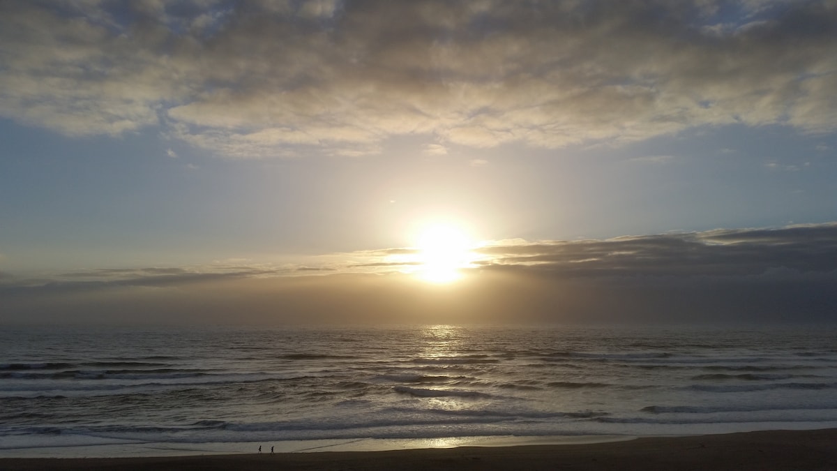 A serene ocean view is captured at sunset, featuring a golden sun dipping below the horizon. Gentle waves roll onto the shore, while the sky is adorned with soft clouds reflecting warm hues.