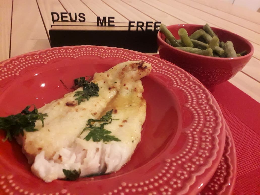 A plate of freshly cooked fish is garnished with herbs, served on a red decorative plate. A bowl of green beans accompanies the dish, sitting beside an inspirational sign that reads 'DEUS ME FREE'. The table setting features neutral tones and a subtle design.
