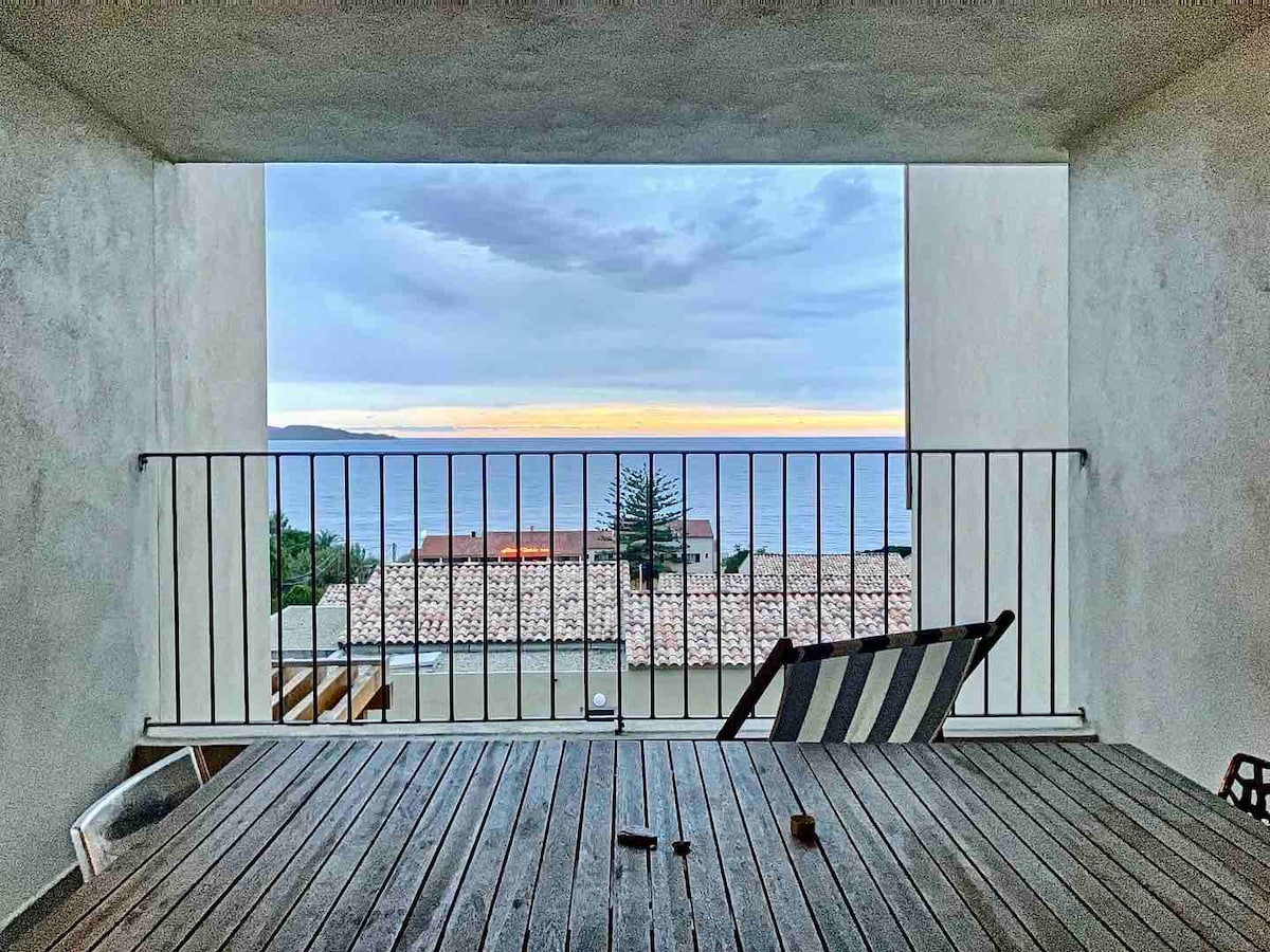 A spacious terrace is featured, surrounded by minimalist architecture. A wooden table is set with a simple design. The view highlights the serene sea horizon, with soft hues of orange and blue during sunset visible through the wide opening. Nearby rooftops are faintly seen.