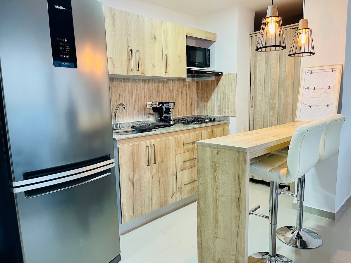 A modern kitchen features wooden cabinetry paired with stainless steel appliances, including a refrigerator and a microwave. A gas stove is set against the back wall, while a light wooden breakfast bar with high stools creates an inviting dining space.
