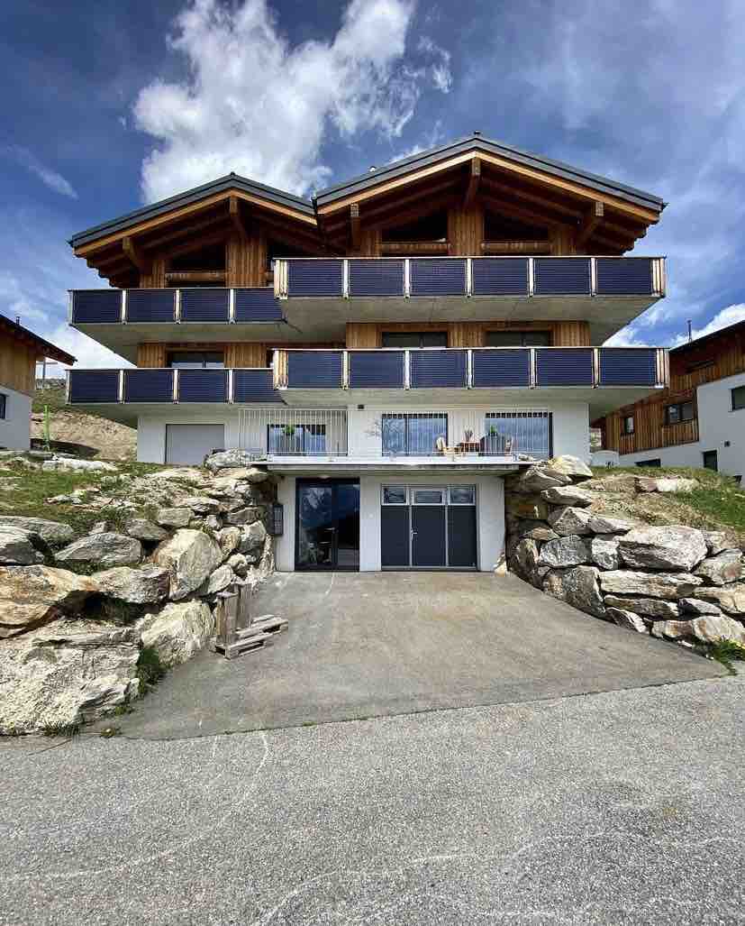 The modern chalet-style building features multiple balconies with wooden accents, set against a backdrop of a clear blue sky. The entrance is framed by natural stone landscaping, providing a welcoming approach to the residence. Ample outdoor space and inviting architecture are on display.