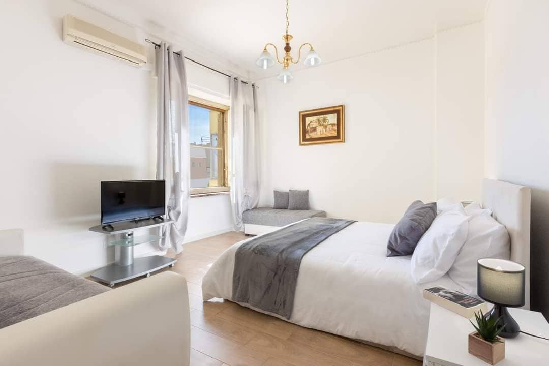 A bright and airy bedroom features a spacious bed dressed in soft linens. A small television is positioned on a stand, while a cozy seating area is visible by the window. Natural light illuminates the room, enhanced by elegant light fixtures.