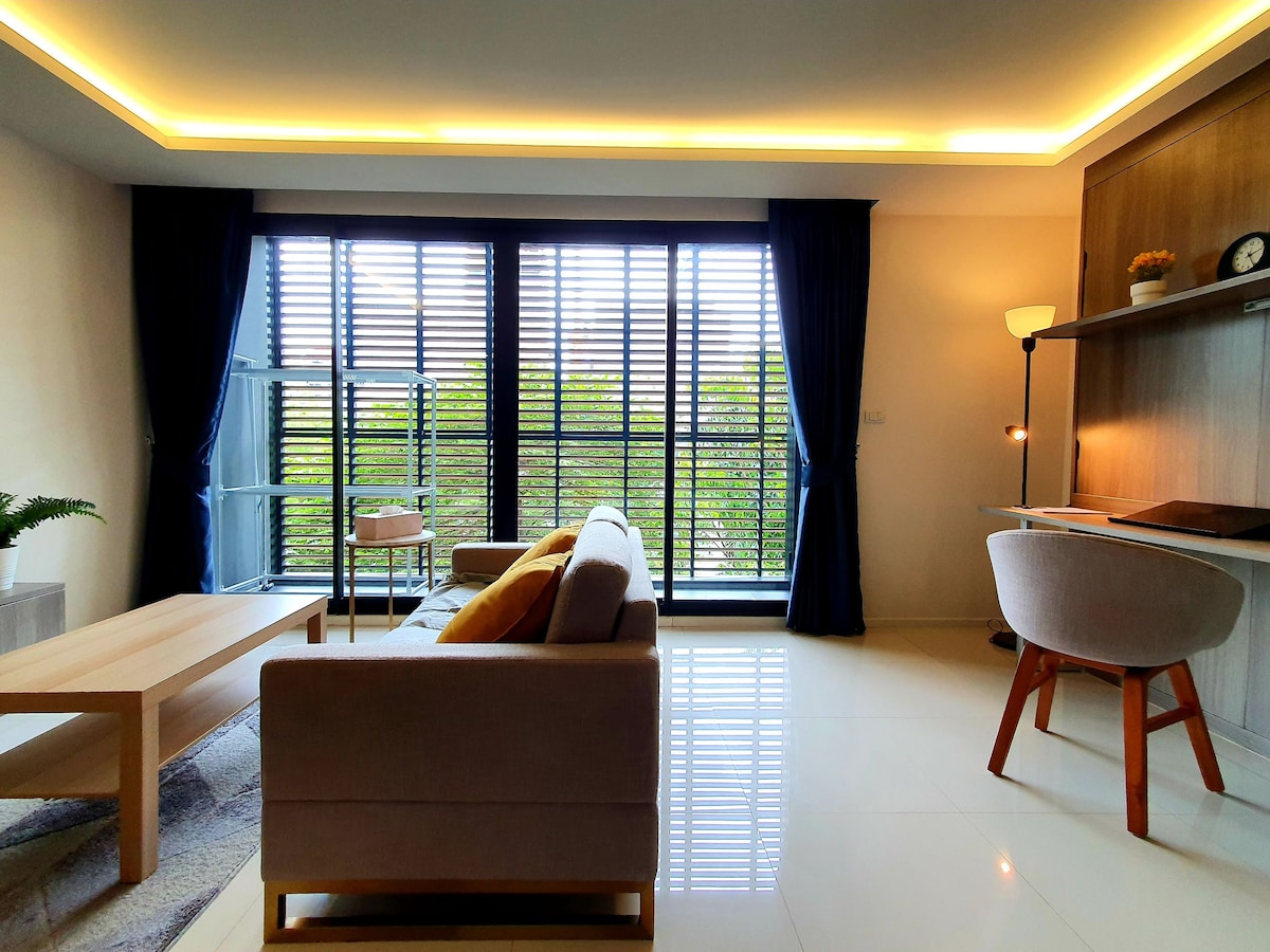 A spacious living area is highlighted by large windows covered with modern slatted blinds, allowing natural light to illuminate the room. A comfortable sofa is complemented by a coffee table and a stylish desk setup. A view of greenery is visible through the windows.