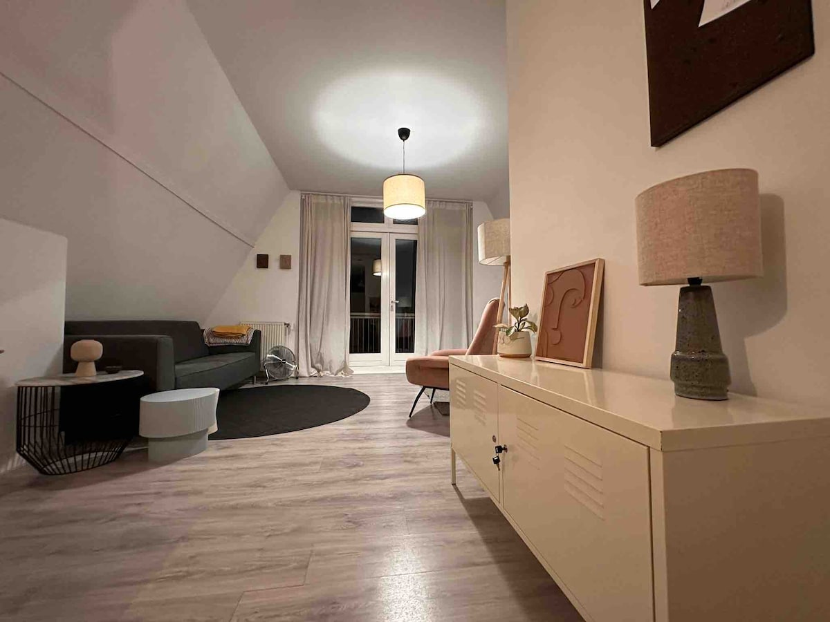 A bright living area features soft lighting from a pendant lamp and side table lamps. A modern sofa is complemented by an accent chair, and a round rug anchors the space. Large windows are adorned with light curtains, allowing natural light to fill the room.