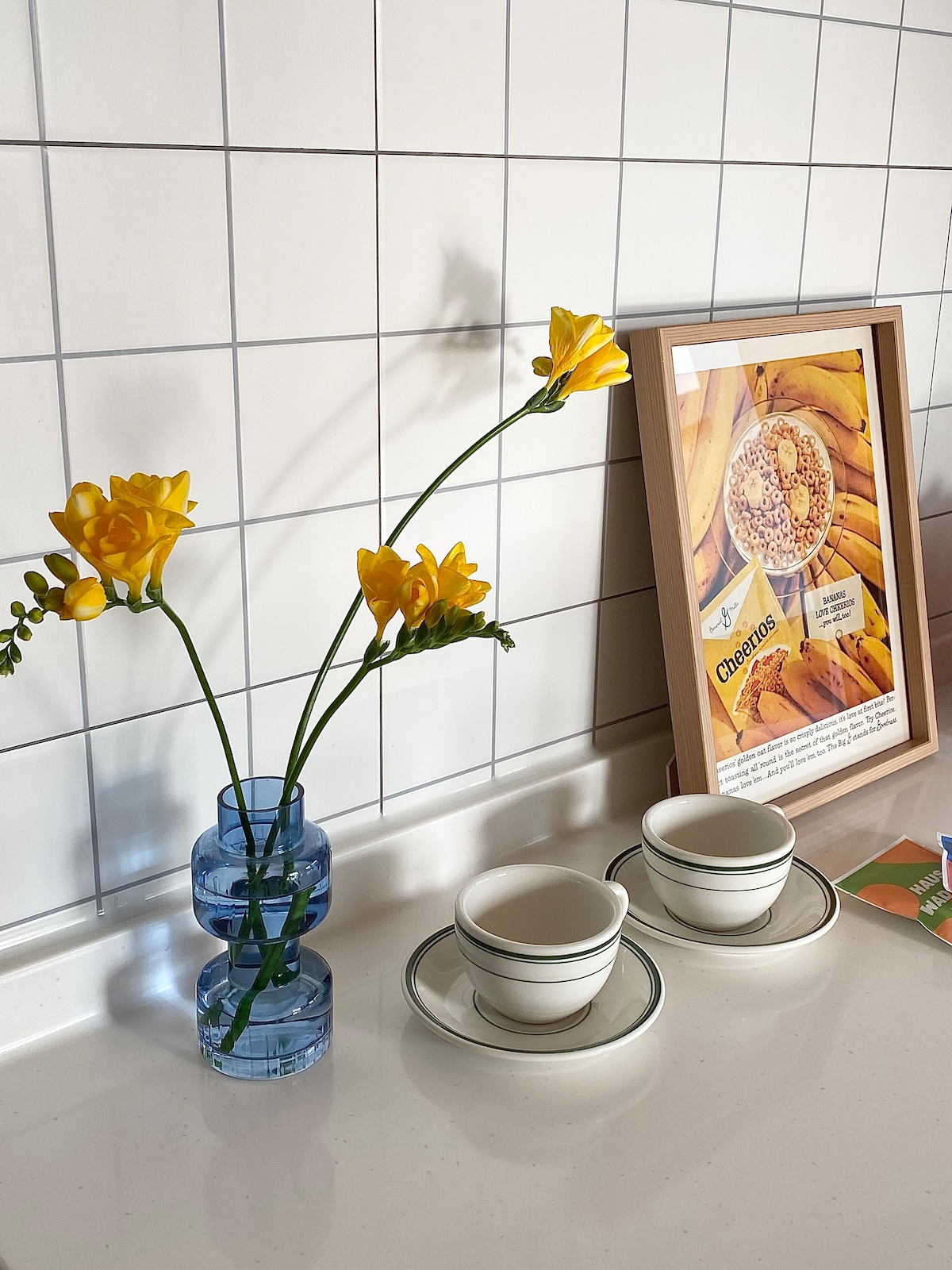 A small table area is highlighted by a blue glass vase holding yellow flowers. Two white cups are positioned on saucers beside a framed print featuring cereal and bananas. The background consists of a grid-patterned tile wall.