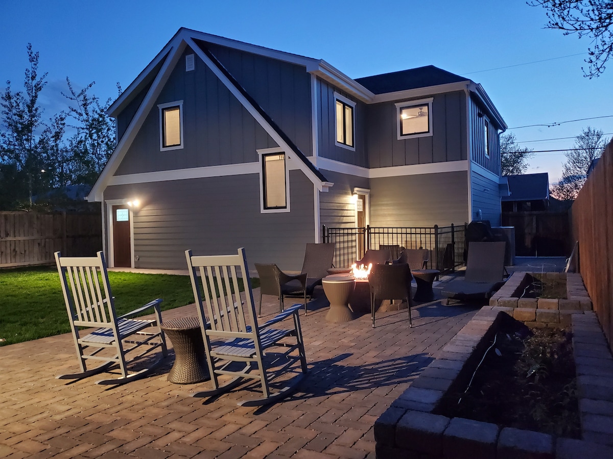 A spacious backyard is highlighted in the evening light, featuring a paved patio area with several seating options, including rocking chairs and a fire pit. The modern house exterior is visible, with large windows reflecting the night sky.