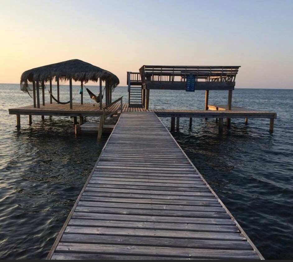 A wooden dock extends over calm waters, leading to two structures. One features a thatched roof and a hammock, while the other provides an open area for relaxation. The horizon showcases a gentle sunset, creating a serene atmosphere.