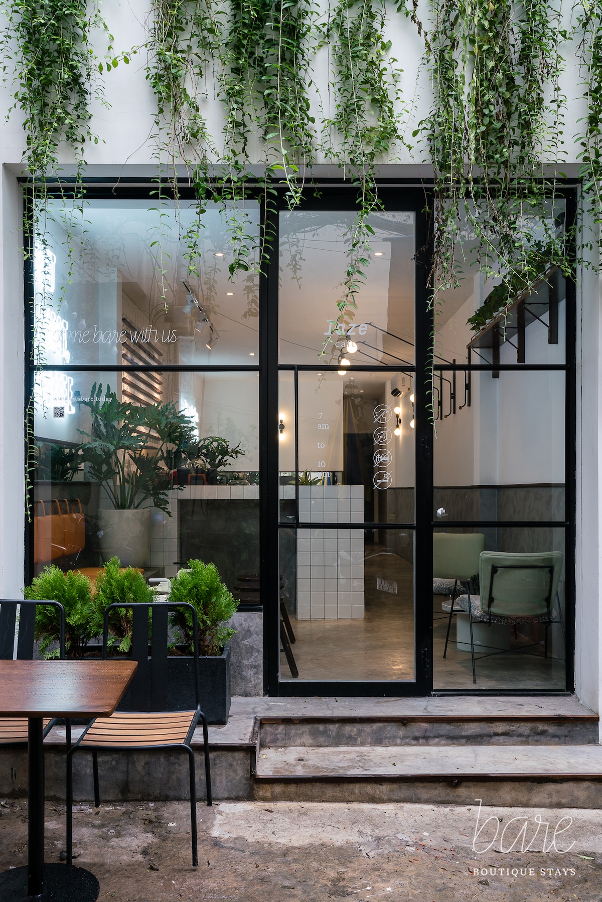 A modern facade features large glass doors framed by lush green plants. Inside, a bright and inviting space is visible, complete with stylish seating and contemporary lighting. The contrasting textures of concrete and greenery create a calm atmosphere that welcomes guests.