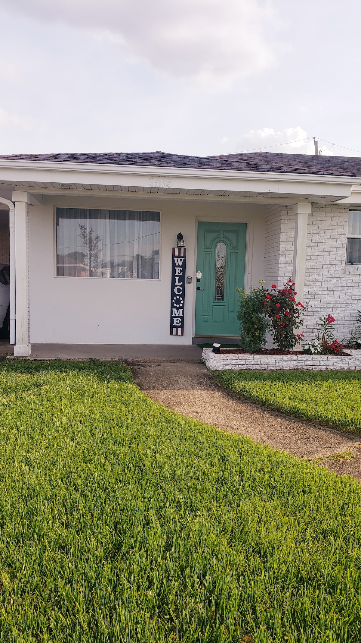 A welcoming entrance features a green door surrounded by white brick, with a decorative sign that reads 'WELCOME.' Vibrant rose bushes are planted beside the porch, and a neatly manicured lawn extends towards the sidewalk.