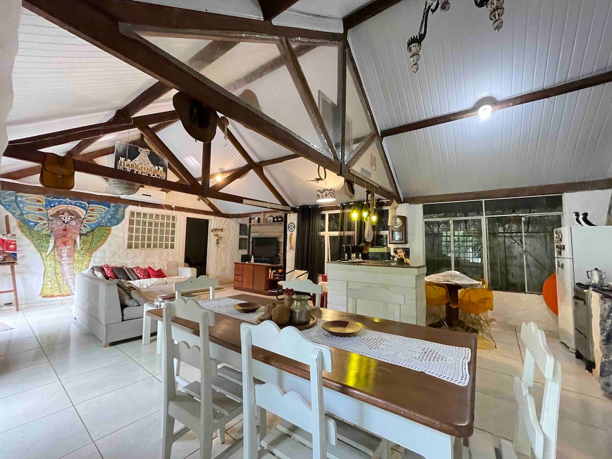 An open living space features a dining area with a wooden table surrounded by white chairs, alongside a comfortable seating area adorned with colorful cushions. Exposed wooden beams frame the high ceiling, while artistic wall decor adds character. Large windows offer a view into the garden.