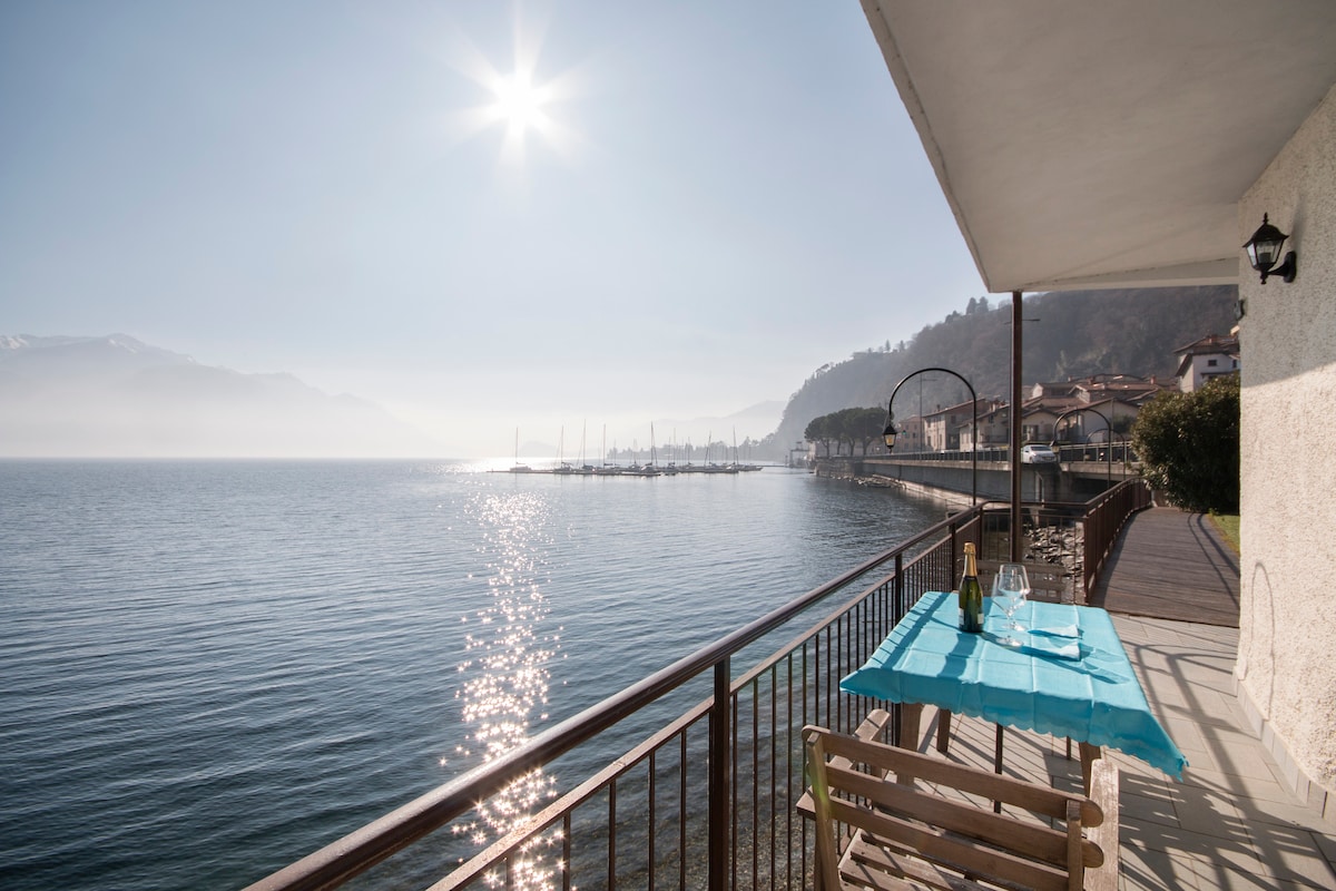 A spacious terrace overlooks the tranquil waters of Lake Como, illuminated by sunlight. A small table with a blue tablecloth is set for two, offering a serene spot to enjoy the view along the sparkling surface of the lake.