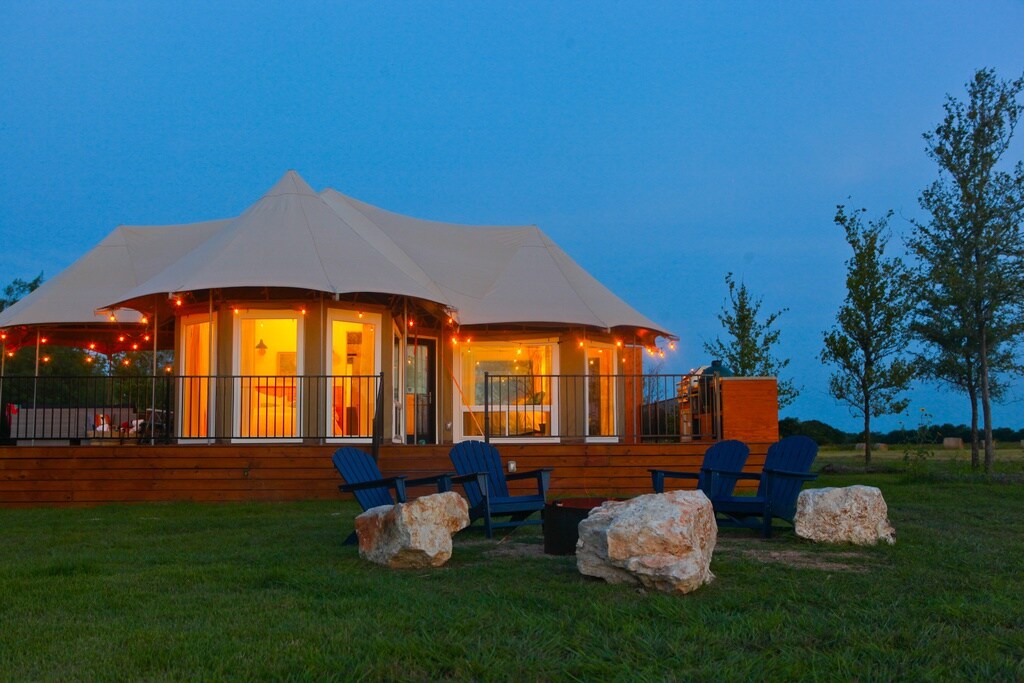 A spacious glamping tent is set against a twilight sky, featuring large windows that glow warmly from within. Outdoor seating is arranged around a fire pit, accompanied by natural stone accents. Lush green grass surrounds the tent, which is framed by trees in the background.