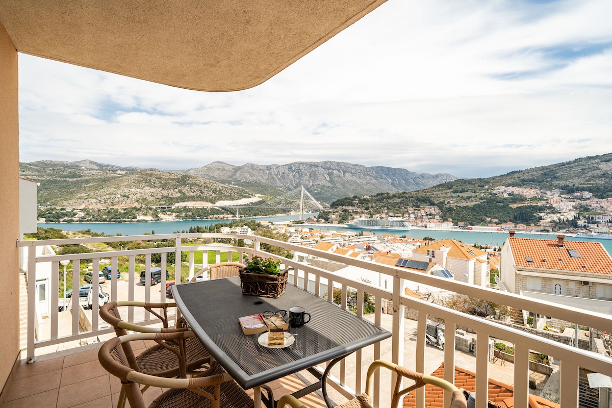 A balcony features a table set for two, complete with coffee mugs and a pastry. The panoramic view showcases the surrounding hills and coastline, with rooftops visible below. The space is enhanced by a potted plant, creating an inviting outdoor seating area.