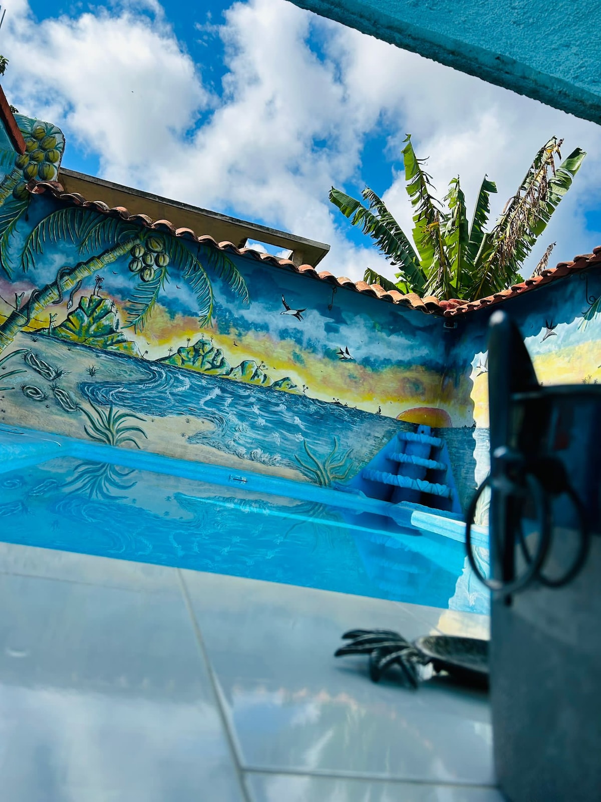 The image depicts a refreshing swimming pool surrounded by vibrant murals depicting tropical scenery. Clear blue water reflects the bright sky and is complemented by the lush greenery of palm trees. A set of steps leads into the pool, inviting relaxation and enjoyment.