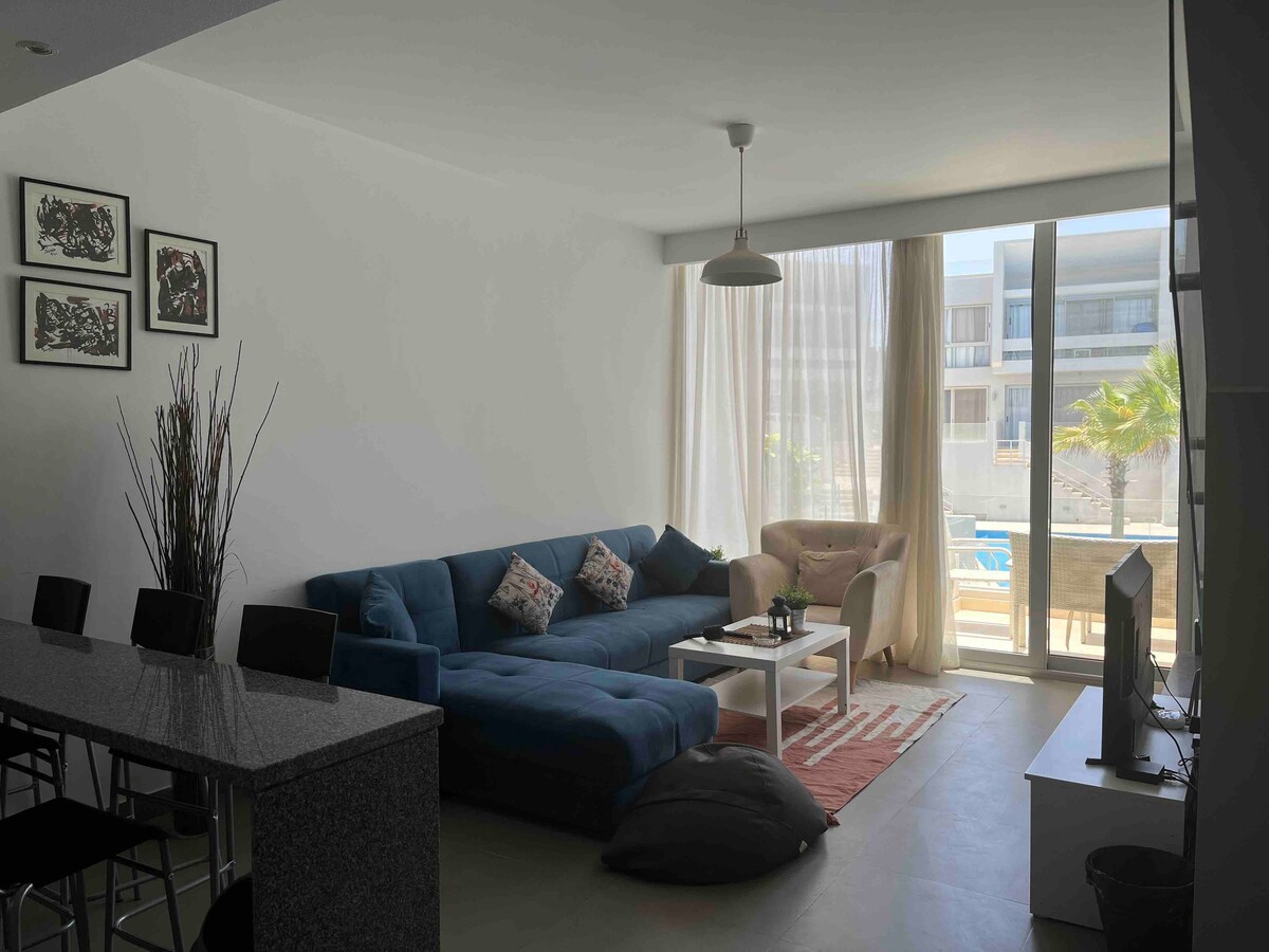A minimalist living room showcases a blue sectional sofa with decorative cushions, paired with a light armchair. Natural light fills the space through large glass doors, leading to a view of the outdoors. A small coffee table sits on a patterned rug, with a television visible nearby.