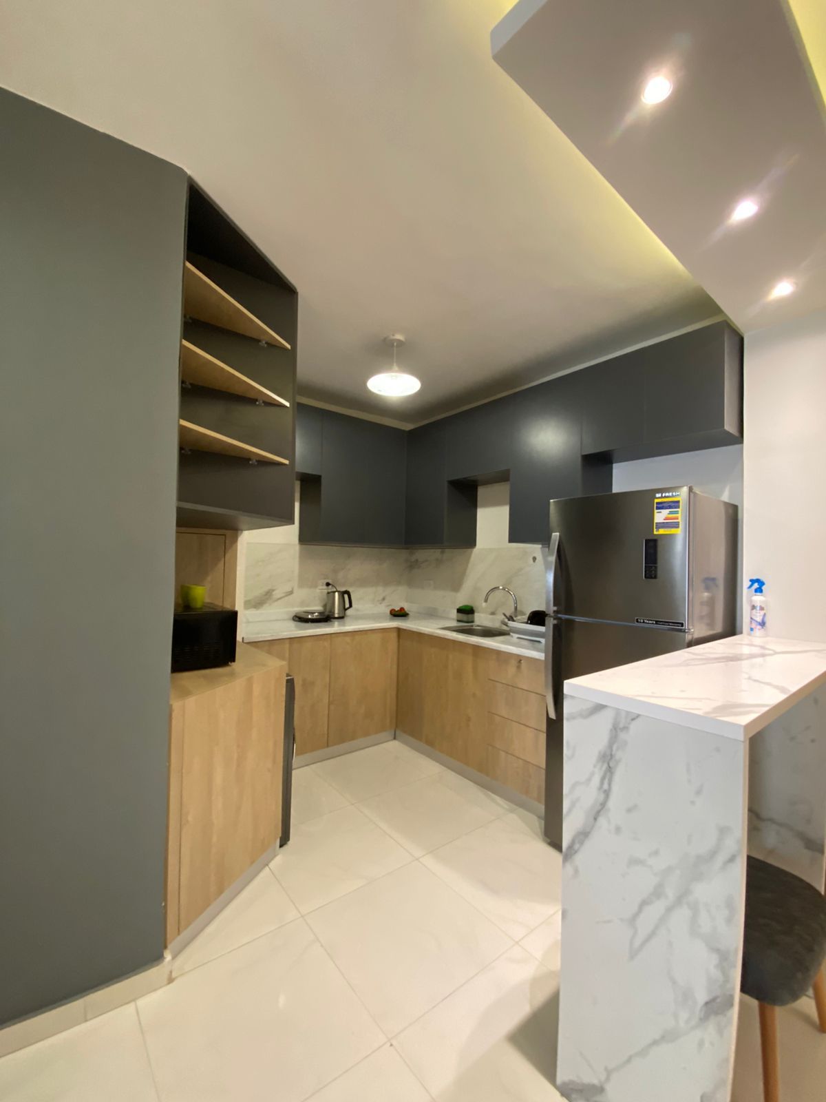 The kitchen features modern cabinetry in grey and wood tones, complemented by a marble countertop. A silver refrigerator stands alongside essential appliances, including a kettle and stovetop, arranged in a functional layout. Shelving above offers extra storage space for kitchen essentials.
