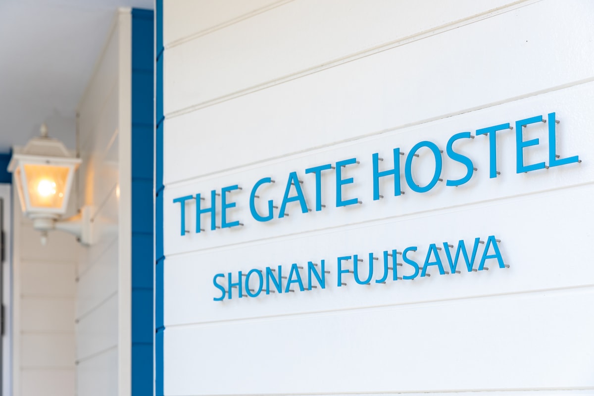 The exterior of the building displays the hostel's name in bold blue letters against a light-colored wall. A vintage lantern hangs nearby, contributing to the building's welcoming appearance.