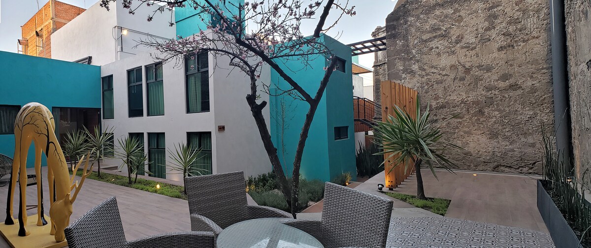 A serene outdoor space features a blend of modern architecture and landscaped areas. A stylized sculpture complements the seating arrangement, which is framed by plants and trees. Natural materials highlight the proximity to the walls of the surrounding buildings, enhancing the private atmosphere.
