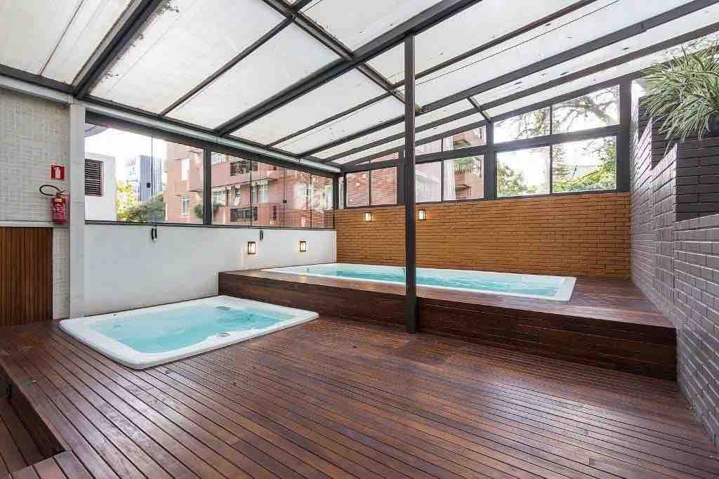 An outdoor area features two hot tubs set on wooden decking, surrounded by a blend of brick walls and green plants. A transparent roof allows natural light to fill the space, creating a warm and inviting environment.