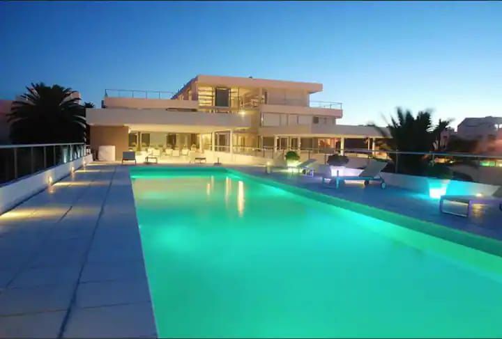 A spacious outdoor area is highlighted by a well-lit pool with a calming turquoise hue. Comfortable lounge chairs are set around the pool, while a contemporary building with large windows provides a backdrop against the evening sky.