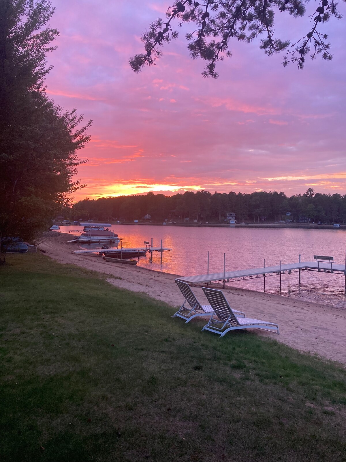 A sandy beach is framed by lush greenery, with two lounge chairs positioned on the grass. A wooden dock extends into the calm lake, where boats are moored. The sunset casts warm hues across the sky, reflecting on the water's surface.